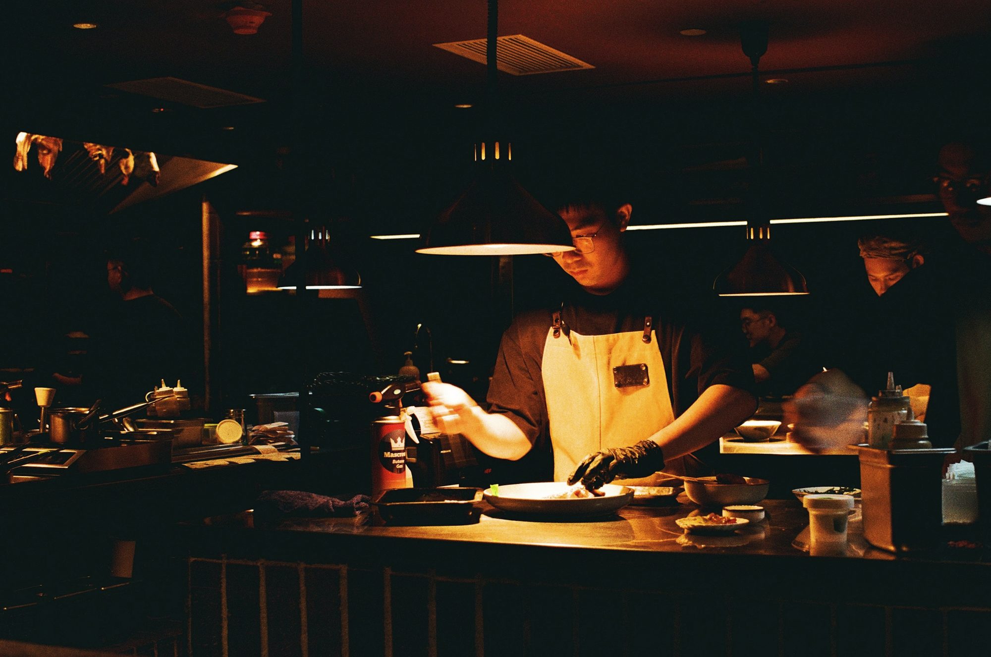 A chef plating under the pass lamps in a dim kitchen